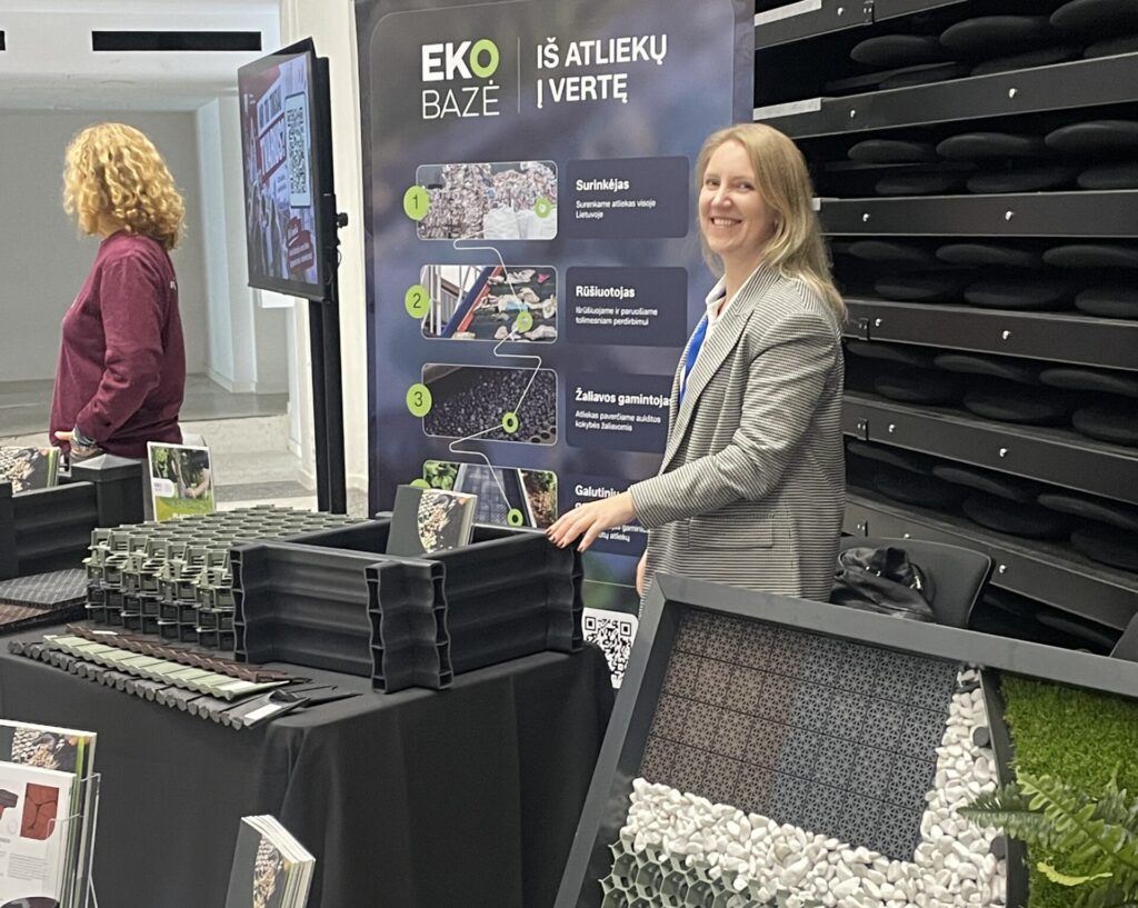 Ekobazė kolegė stovi prie stendo ir rodo į galutinius gaminius pagamintus iš perdirbto plastiko. Jaunimo tvarumo festivalis 2926 Litexpo