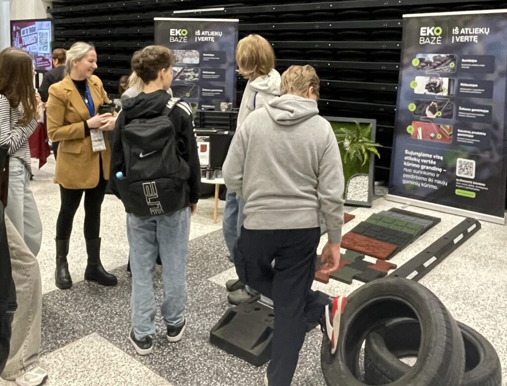 Mokiniai susidomėję klausosi Ekobazė kolegės, stovinčios prie stendo ir laikančios žaliavų pavyzdžius rankose. Jaunimo tvarumo festivalis 2926 Litexpo